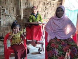 A mother and her two daughters pose for the picture, in the foreground on the left a little girl with a quilt and a yellow top, her mother on the right and in the background her older daughter in a wheelchair. They are inside their shelter.