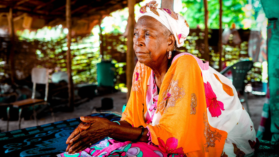 Portrait of an elderly woman dressed in a colorful dress and scarf, seated in the shade of a straw hut. She is looking to the left, holding her hands in her lap.