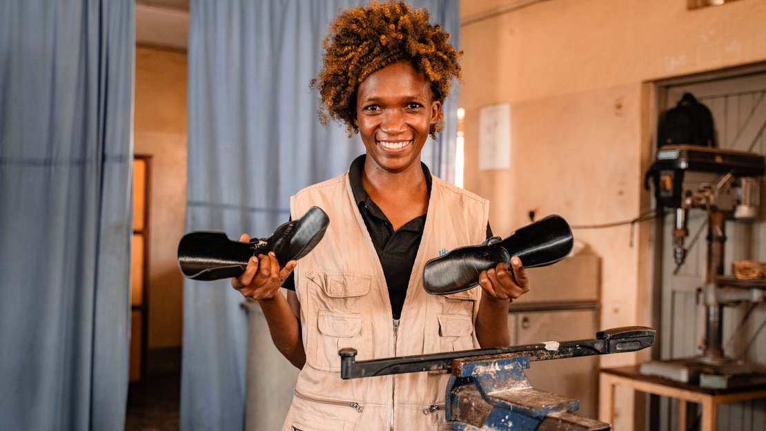 A young black woman with curly hair is in a brace shop. She holds a black plastic brace in each hand and smiles at the camera.