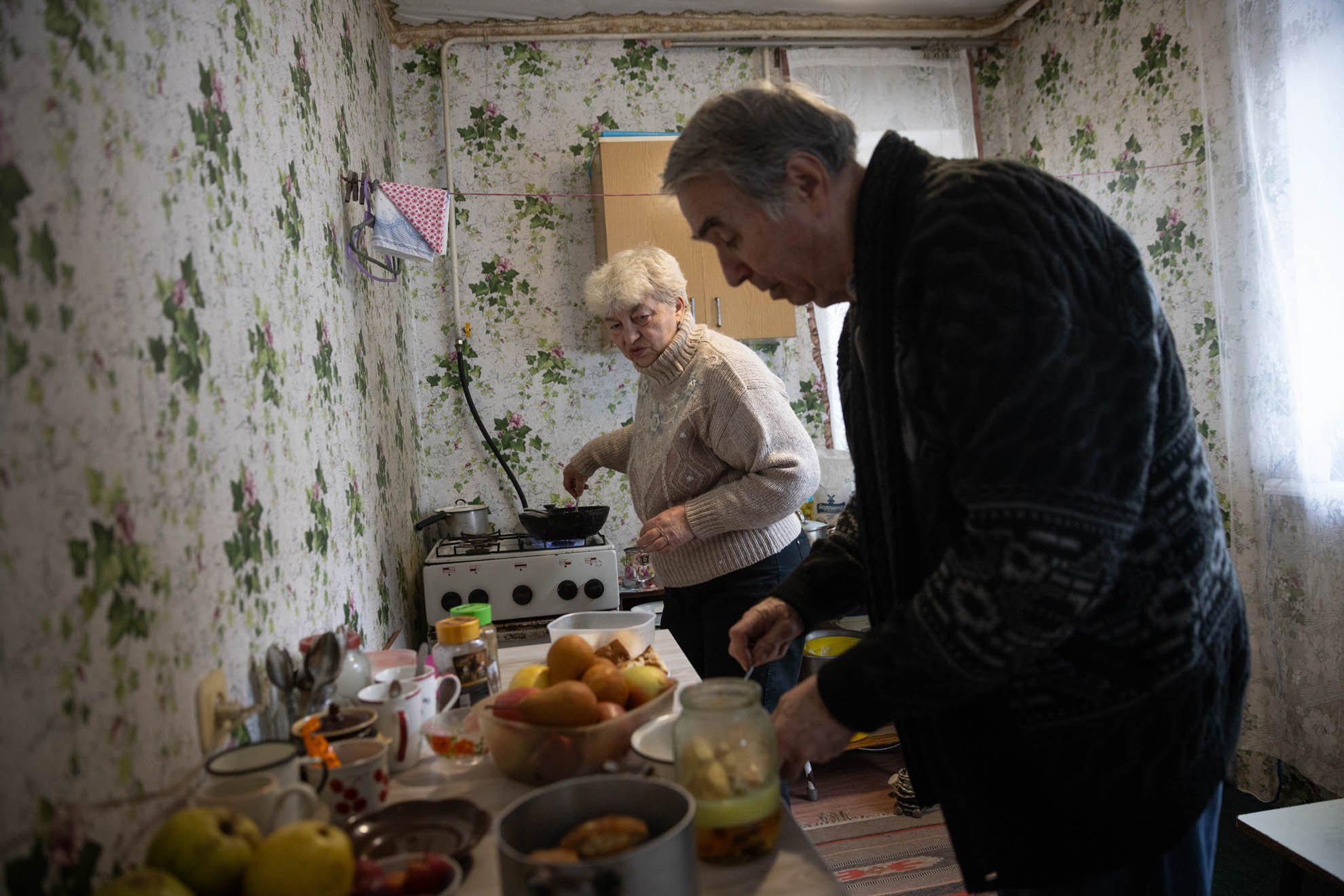 Yevhen’s parents are cooking in their kitchen that has flowery wallpaper. 