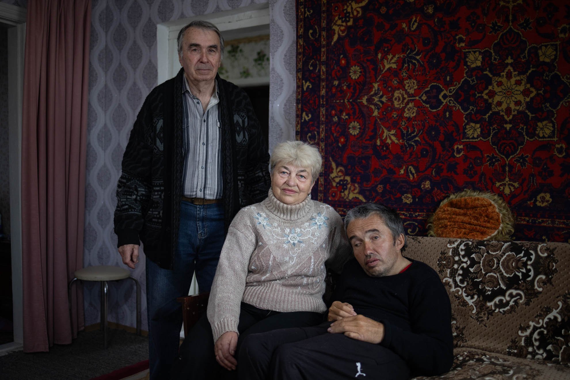 Yevhen is sitting on a brown sofa with his mother sitting next to him and his father standing behind them. 